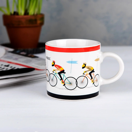 White mug with cyclist design on a table with a plant and remote control in the background
